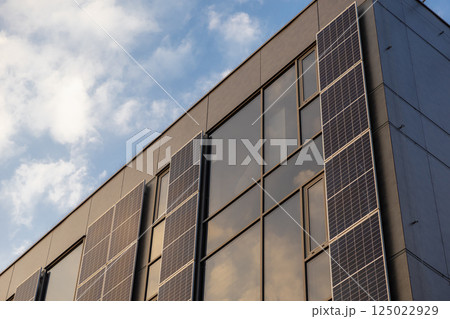 Modern building facade with vertical solar panels and large glass windows reflecting soft evening sky 125022929