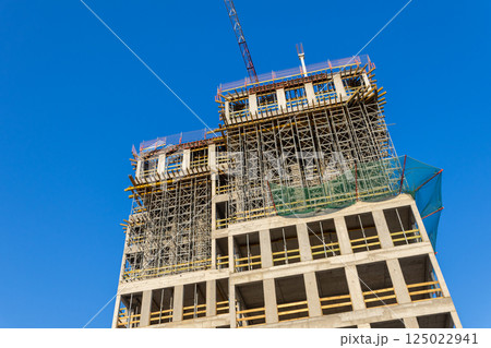 Modern high-rise building under construction with scaffolding and concrete structure against clear blue sky during daytime urban development process 125022941
