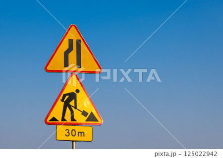Road construction and lane reduction warning signs against clear blue sky indicating roadwork ahead in 30 meters distance on highway or city road Road construction and lane reduction warning signs against clear blue sky indicating roadwork ahead in 30 meters distance on highway or city road 125022942
