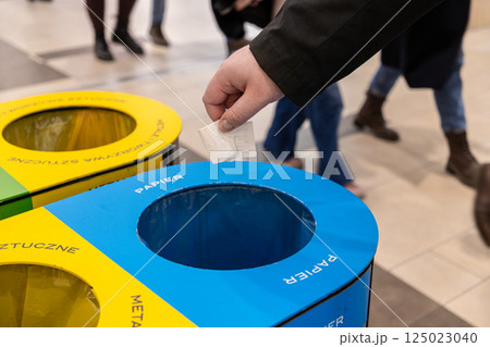 Person throwing paper receipt into blue recycling bin labeled papier in shopping mall, with colorful containers for plastic and metal in background Person throwing paper receipt into blue recycling bin labeled papier in shopping mall, with colorful containers for plastic and metal in background 125023040