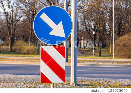 road signs with blue direction arrow and red warning stripes on metal poles near empty street and trees in background during sunny day in urban environment 125023045