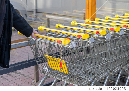 Row of shopping carts with yellow handles connected by coin chain locks near glass wall outside supermarket entrance with hand grabbing one trolley 125023068