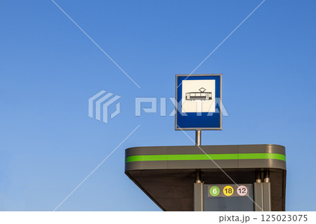 Tram stop sign on top of modern shelter against clear blue sky with line numbers visible and no people around on sunny day in urban public transportation area Tram stop sign on top of modern shelter against clear blue sky with line numbers visible and no people around on sunny day in urban public transportation area 125023075