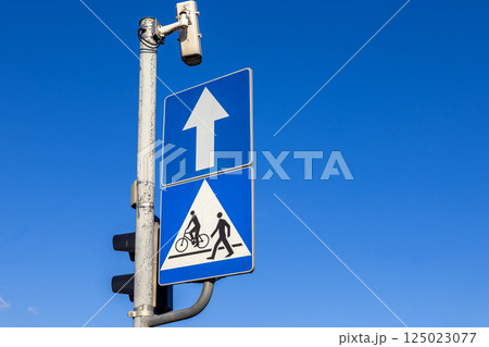 Traffic signs with straight ahead arrow and pedestrian crossing with cyclist symbol under blue sky near mounted surveillance camera Traffic signs with straight ahead arrow and pedestrian crossing with cyclist symbol under blue sky near mounted surveillance camera 125023077