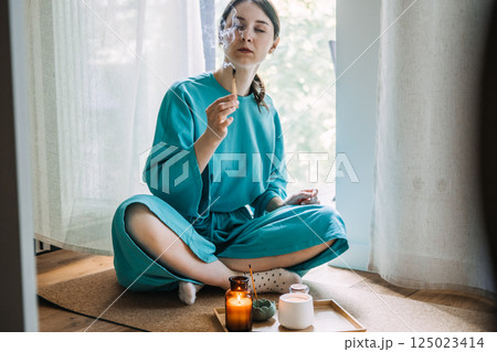 Young woman holding burning palo santo while seated cross-legged beside lit candles and incense. Home sanctuary and personal zen zones, palo santo ritual, curated calm, domestic tranquility 125023414