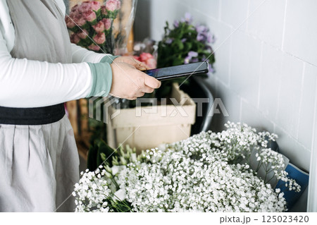 Modern florist using a smartphone while working with baby's breath and flower inventory. Floral content creation, smartphone photography, botanical influencers, modern florists 125023420