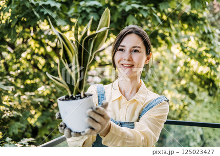 Young Asian woman smiling while holding a potted snake plant toward the camera on a leafy balcony. Everyday wellness, tactile moments, plant care, grounded joy 125023427