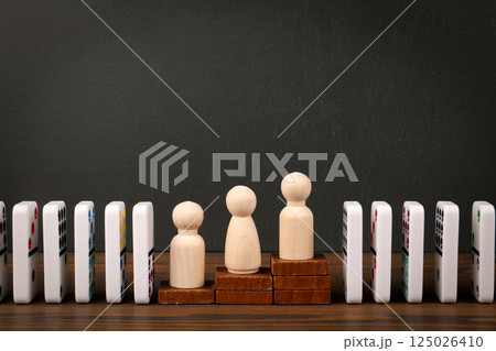 Wooden Figures Standing on Stacked Blocks with Dominoes in Background Wooden Figures Standing on Stacked Blocks with Dominoes in Background 125026410