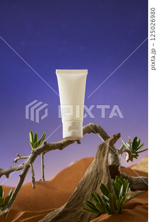 Vertical frame of unlabeled cosmetic tube placed on a crooked dry tree branch. The dry branch has a grayish-brown color. Green leaves with roots buried in sand grow lushly. Desert concept.  125026980