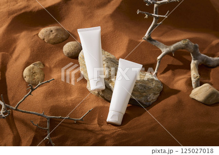 Top view of cream tubes placed on a light colored stone slab with a rough surface and dark veins. Pebbles are scattered around. Barren tree branches with many sharp thorns are displayed on the sand. 125027018