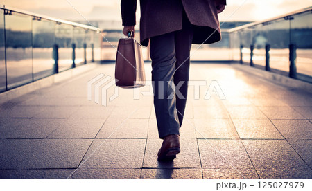 a businessman walking with a leather briefcase in hand on a modern pedestrian walkway during sunrise, symbolizing business travel, career success, leadership, and future goals, corporate, finance 125027979