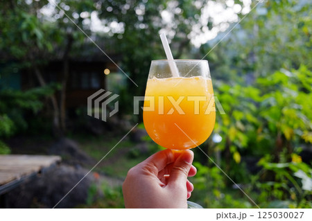 Hand holding a single glass of ice orange juice in natural garden of cafe in tropical forest Hand holding a single glass of ice orange juice in natural garden of cafe in tropical forest 125030027