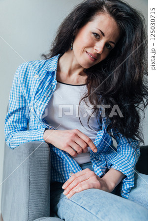 brunette woman in a blue shirt sitting on an armchair and smiling 125030715