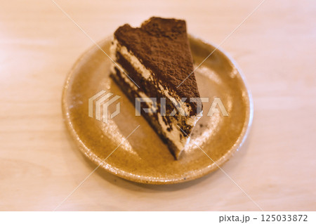 Slice of delicious tiramisu cake on ceramic plate served on wooden table in warm cozy cafe atmosphere Slice of delicious tiramisu cake on ceramic plate served on wooden table in warm cozy cafe atmosphere 125033872