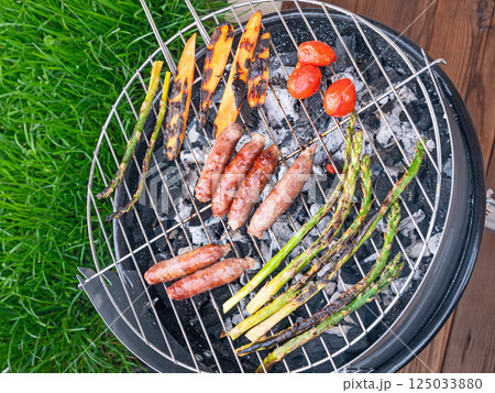 close-up top view of a charcoal grill with sausages, asparagus and cherry tomatoes being grilled outdoors at backyard 125033880