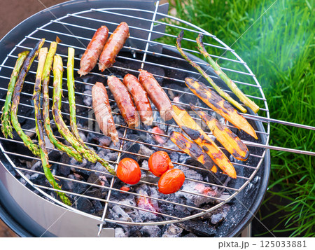 close-up top view of a charcoal grill with sausages, asparagus and cherry tomatoes being grilled outdoors at backyard 125033881