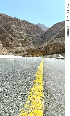 Ras Al Khaimah,UAE - March 23, 2025:Curved highway winds gracefully between canyon cliffs, road alignment flows like ribbon, sharp rocks cast strong shadows as travelers follow path into raw Ras Al Khaimah,UAE - March 23, 2025:Curved highway winds gracefully between canyon cliffs, road alignment flows like ribbon, sharp rocks cast strong shadows as travelers follow path into raw 125034103