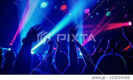 Energetic crowd dancing and raising hands at a live music concert, illuminated by vibrant laser lights, creating a captivating slow motion spectacle 125034108