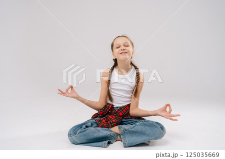 Serene child gel meditating sitting with closed eyes in lotus pose, performing mudra hand gesture in studio with white background showcasing mindfulness and tranquil inner state. 125035669