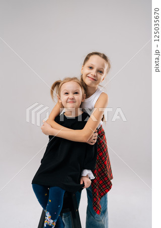 Joyful two young sisters posing for studio portrait, with older sister affectionately hugging younger sister from behind, both smiling happily looking at camera. Concept of happy sisterhood and family 125035670