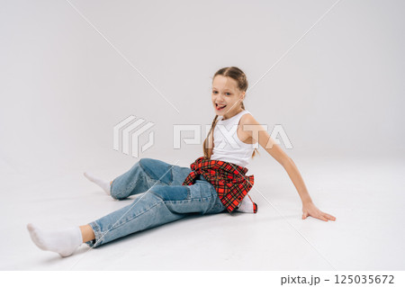 Front view of cheerful preteen girl with braided hair, playfully sticking out tongue while sitting on floor in studio with white isolated background. Concept of carefree childhood. 125035672