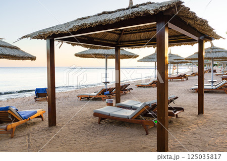 Relaxing beachside cabanas provide shade at sunset on a serene shoreline with lounge chairs Relaxing beachside cabanas provide shade at sunset on a serene shoreline with lounge chairs 125035817