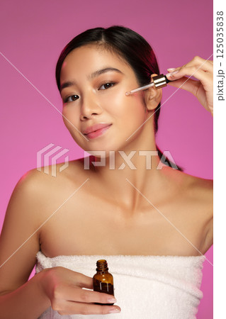 Woman applying facial serum with a dropper while smiling against a vibrant pink background Woman applying facial serum with a dropper while smiling against a vibrant pink background 125035838
