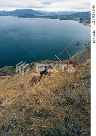 Hiking down a steep hillside towards a serene lake with mountains in the distance during a cloudy day Hiking down a steep hillside towards a serene lake with mountains in the distance during a cloudy day 125035846
