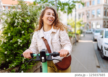 Happy woman rides an electric scooter in the city street on a sunny day. Ecological transport. Happy woman rides an electric scooter in the city street on a sunny day. Ecological transport. 125037885
