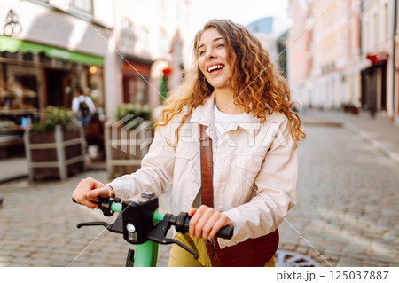 Happy woman rides an electric scooter in the city street on a sunny day. Ecological transport. Happy woman rides an electric scooter in the city street on a sunny day. Ecological transport. 125037887