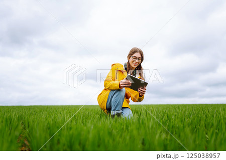Young farmer woman in a green field, intently using a tablet. She wears a yellow jacket and glasses Young farmer woman in a green field, intently using a tablet. She wears a yellow jacket and glasses 125038957