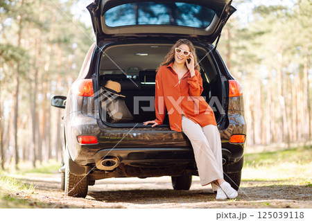 A traveler young woman smiles while sitting in the trunk of her car during a sunny day in a forest A traveler young woman smiles while sitting in the trunk of her car during a sunny day in a forest 125039118