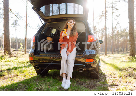 Beautiful woman sits in the trunk of a car outdoor and uses a smartphone. Lifestyle 125039122