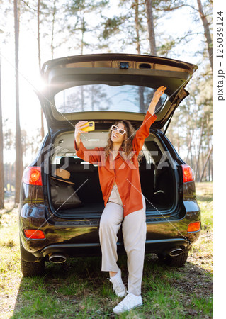 Happy woman sits on the trunk of her car and taking a selfie. Selfie while traveling by car. 125039124