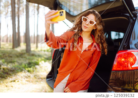 Happy woman sits on the trunk of her car and taking a selfie. Selfie while traveling by car. 125039132