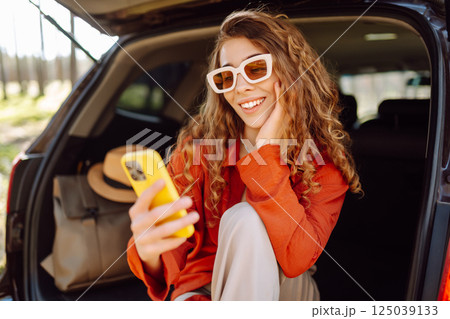 Beautiful woman sits in the trunk of a car outdoor and uses a smartphone. Lifestyle 125039133