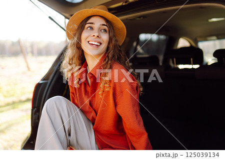 A traveler young woman smiles while sitting in the trunk of her car during a sunny day in a forest 125039134