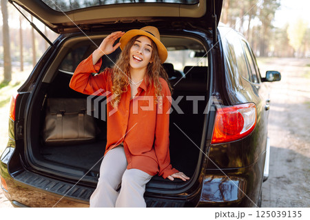 A traveler young woman smiles while sitting in the trunk of her car during a sunny day in a forest 125039135
