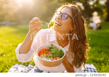 Woman enjoying a poke bowl on sunny day in a park while sitting on a picnic blanket. Concept picnic Woman enjoying a poke bowl on sunny day in a park while sitting on a picnic blanket. Concept picnic 125039345