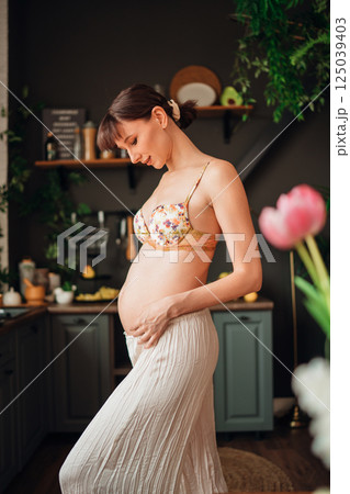 Young pregnant woman gently touching her belly in a cozy kitchen setting Young pregnant woman gently touching her belly in a cozy kitchen setting 125039403