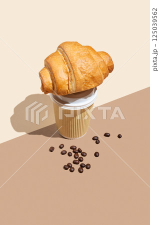 Paper cup with scattered coffee beans and fresh croissant on yellow and brown background. 125039562