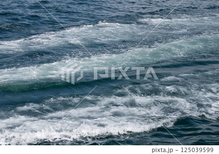 Ocean waves forming white foam trail on deep blue sea surface during daytime. 125039599