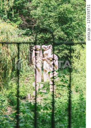 Weathered stone statues of young women figures stand hidden behind lush forest fence. Mysterious scene from folk legend, statues evokes memory, loss, and forgotten stories 125039731