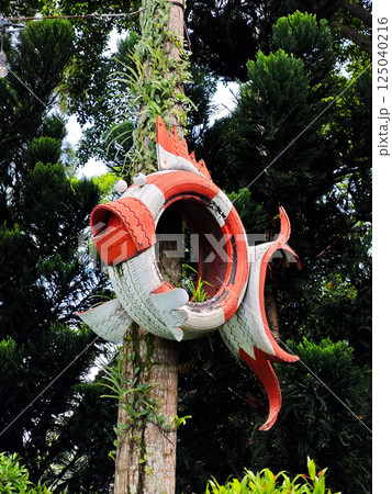 Creative orange tire sculpture resembling a fish attached to a tree in an outdoor garden with a green foliage background 125040216