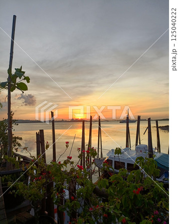 Serene Sunset Over a Calm Waterfront with Boats and Vibrant Flowers at a Coastal Dock in Johor Bahru Malaysia with Industrial Silhouette on the Horizon 125040222