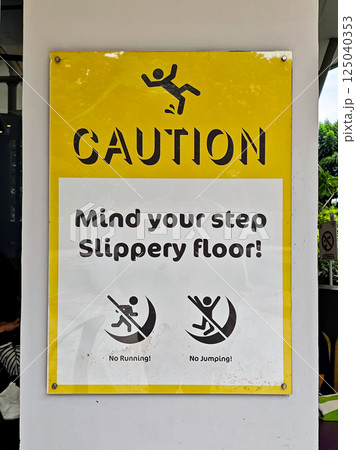 Bright yellow caution sign with stick figure and text "Mind your step Slippery floor" posted on a swimming pool area emphasizing safety precautions and warning against running or jumping. Bright yellow caution sign with stick figure and text "Mind your step Slippery floor" posted on a swimming pool area emphasizing safety precautions and warning against running or jumping. 125040353