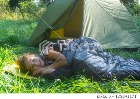 The girl is lying in a sleeping bag on a warm mat near a tent outdoor. Family camping recreation, hiking equipment. The girl is lying in a sleeping bag on a warm mat near a tent outdoor. Family camping recreation, hiking equipment. 125041171