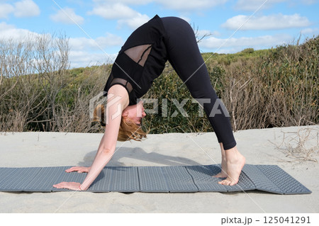 Woman practicing yoga outdoors on sandy surface, performing downward dog pose on a mat, surrounded by nature under blue sky with light clouds, peaceful atmosphere. 125041291