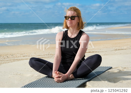 Woman practicing yoga on sandy beach, sitting in butterfly pose on mat, embracing mindfulness, relaxation, and wellness with ocean waves and blue sky background. Woman practicing yoga on sandy beach, sitting in butterfly pose on mat, embracing mindfulness, relaxation, and wellness with ocean waves and blue sky background. 125041292
