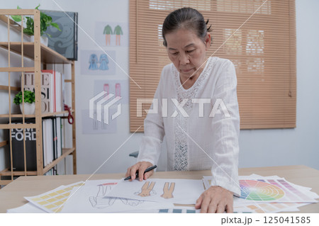 An older woman analyzes creative designs at her desk, showcasing her dedication to art and design in a professional setting. 125041585